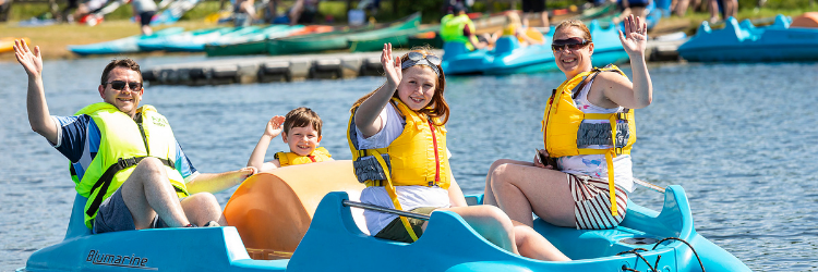 Photograph of a happy family, all smiling and waving at the camera, while riding a pedalo on Dinton lake on a beautiful sunny day