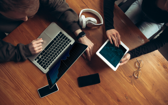 Birds eye view of a table with two people using a laptop and ipad, along with mobile phones and a pair of headphones