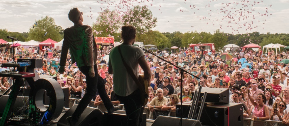 Two young rock musicians, seen from behind, performing on a stage in front of a large crowd as confetti is launched into the audience