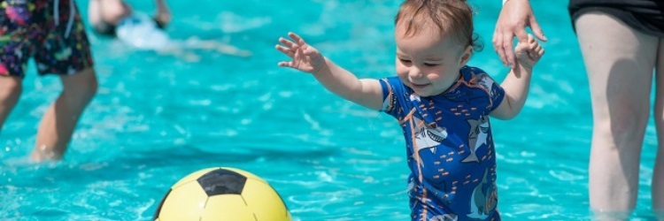 a smiling young child in a paddling pool, holding an adult's hand while looking at a ball floating on the surface