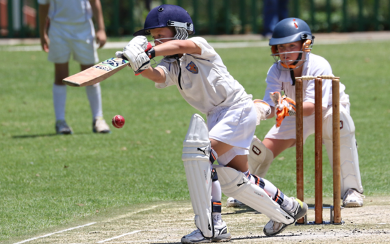 A batsman plays a drive at a cricket ball, with the wicketkeeper watching on