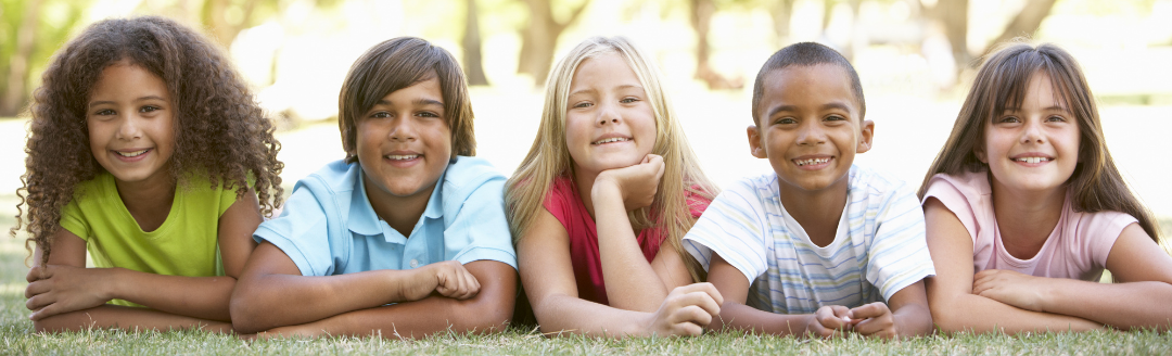 five children, mixed boys and girls, laying front down on the ground outside on a sunny day and looking happily at the camera