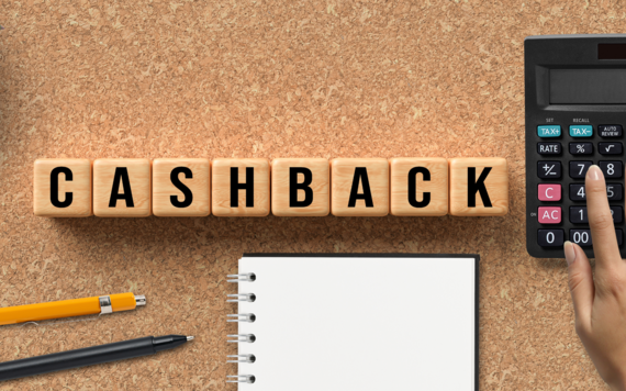 The word cash back in the middle of a cork panel with a hand on a calculator to its right and a note book and two pens underneath