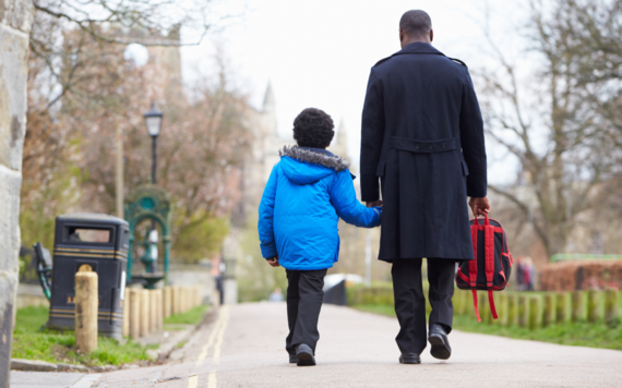 The back view of a father and son walking along a path holding hands with the father carrying the sons school backpack