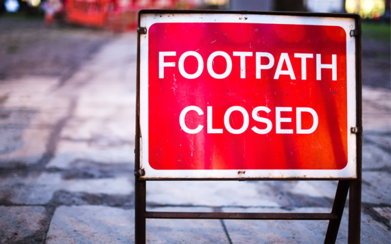 Red sign with white lettering that says footpath closed standing on a pavement