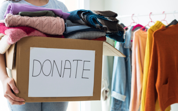 A women holding a box of clothes, with the word donate in the front. Some clothes are hanging on a rail next to her