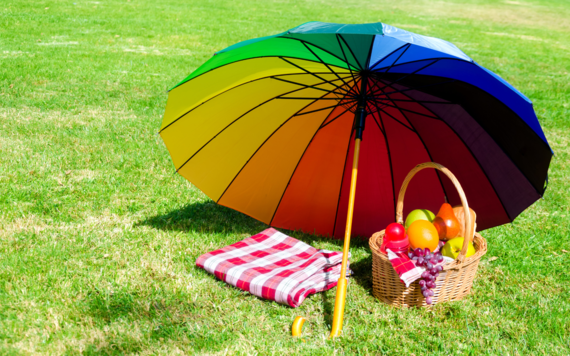 Rainbow umbrella sheltering a picnic basket and blanket from the sun in a park 