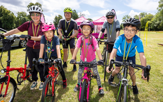 Three children in front and three adults behind them on bikes wearing helmets in a field with a gazebo in the distance behind them.