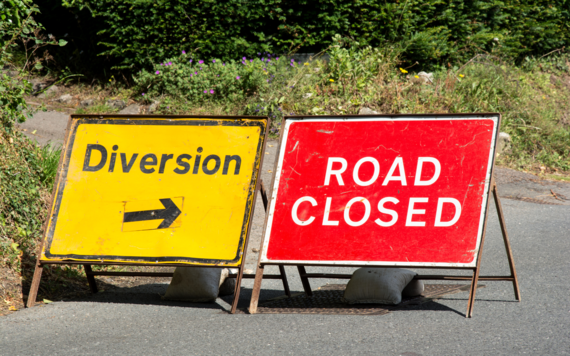 Two signs on a road with hedgerow in the background. On the left a diversion sign with an arrow pointing right, on the right a road closed sign