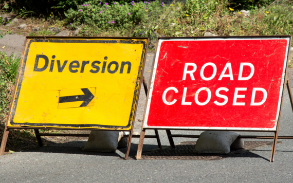Two signs on a road with hedgerow in the background. On the left a diversion sign with an arrow pointing right, on the right a road closed sign