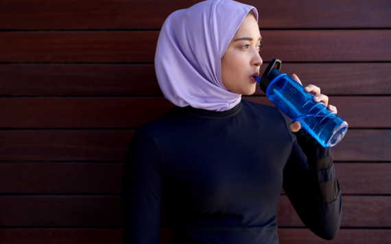 A lady wearing a lilac hijab and black top standing next to a fence drinking a flask of water