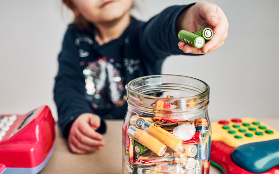 A photo of a boy putting two used batteries in a jar full of batteries. There are two toys on each of his two sides