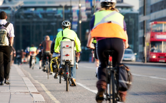 Shot from behind, a line of cyclists in a cycle lane with people walking on the pavement and traffic on the road