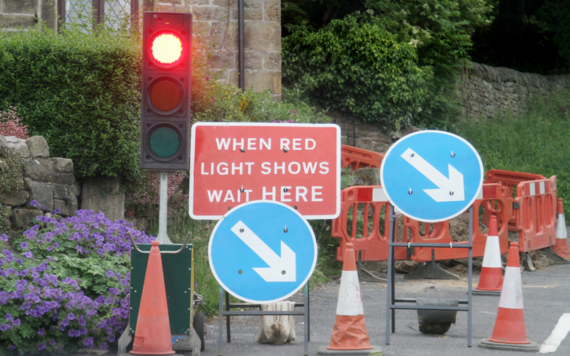 Temporary traffic light on red, a sign that says when red light shows wait here, two signs with arrows in blue circles, some cones and barriers