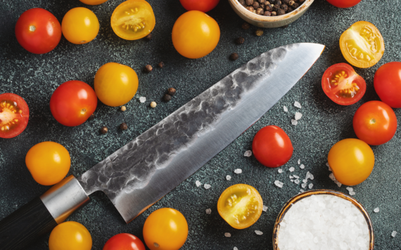 An image of a kitchen knife on a table, with some cherry tomatoes and a bowl of salt and peppercorn around it