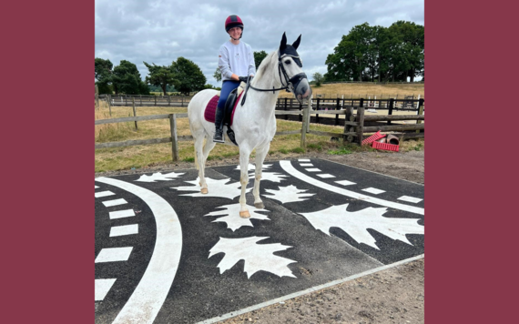 A rider sat on a white horse which is standing on the tarmac decorated with paintings of leaves