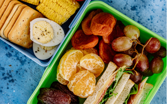 Two lunchboxes. One has sandwiches and a mix of fresh and dry fruit, the other hard boiled eggs, crackers and corn on the cob