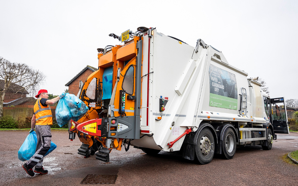 A waste worker loads blue bags into the back of a rubbish truck