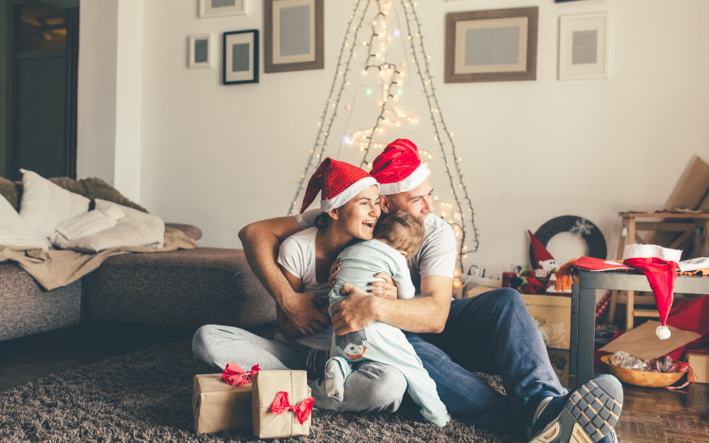 A mother and father in Christmas hat cuddle their child with Christmas tree and presents around them