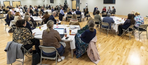 wide shot of a large room filled with people sitting around tables listening to a speaker