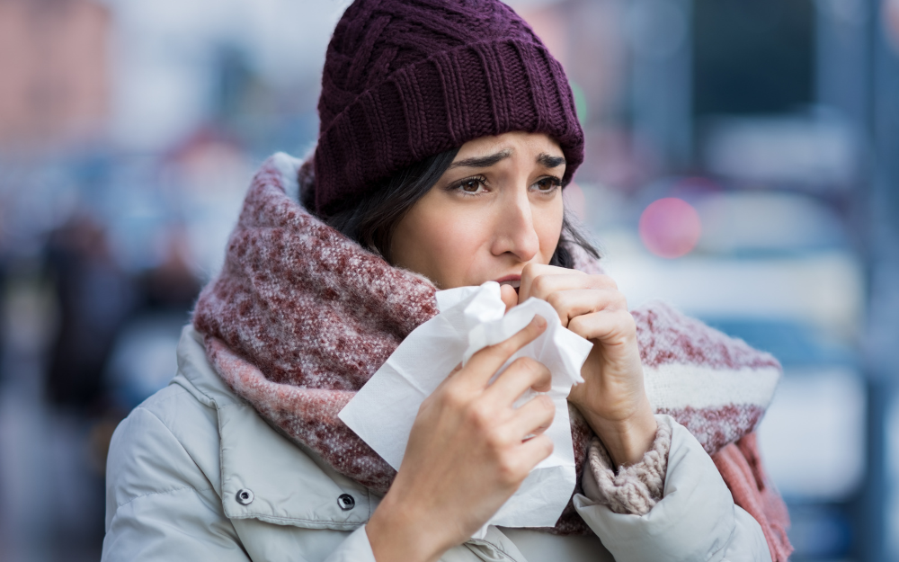 A woman wrapped in hat and scarf holds a tissue as she coughs in the cold air