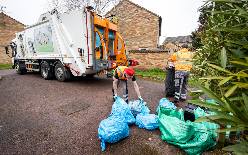 Two men collect rubbish and recycling, loading it from the kerbside into a waste truck
