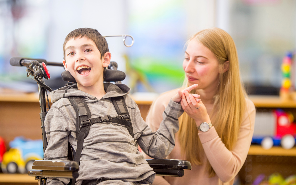 A caregiver helps a young boy with a physical disability. The boy is happily smiling and laughing