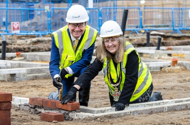 A man and a woman in high-viz jackets and helmets smile while kneeling on the ground and laying the first bricks on a building site
