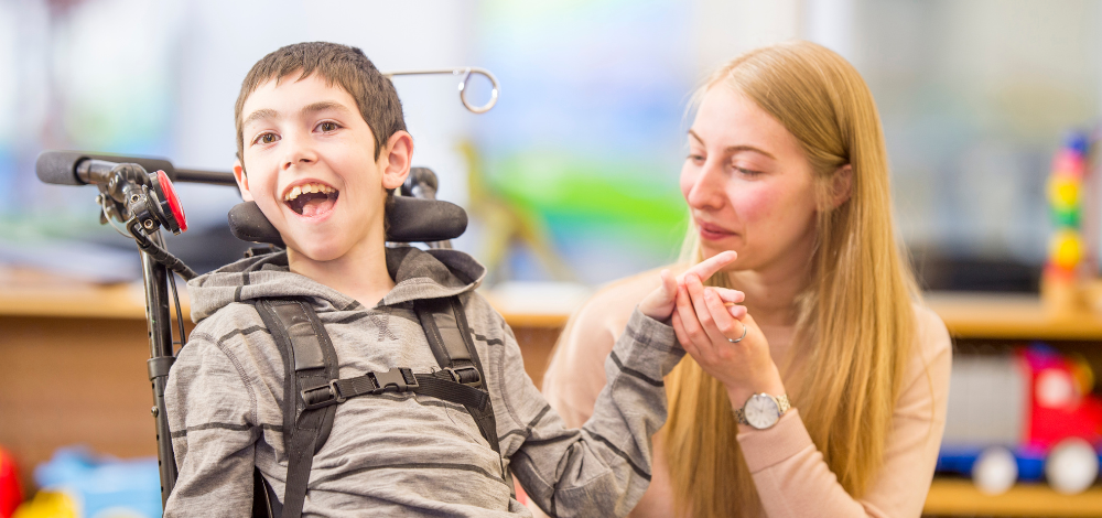 a child in a wheelchair smiles enthusiastically at the camera as an adult holds his hand