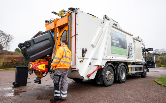 A crew seeing waste in a black wheeled bin being loaded to a waste truck. 
