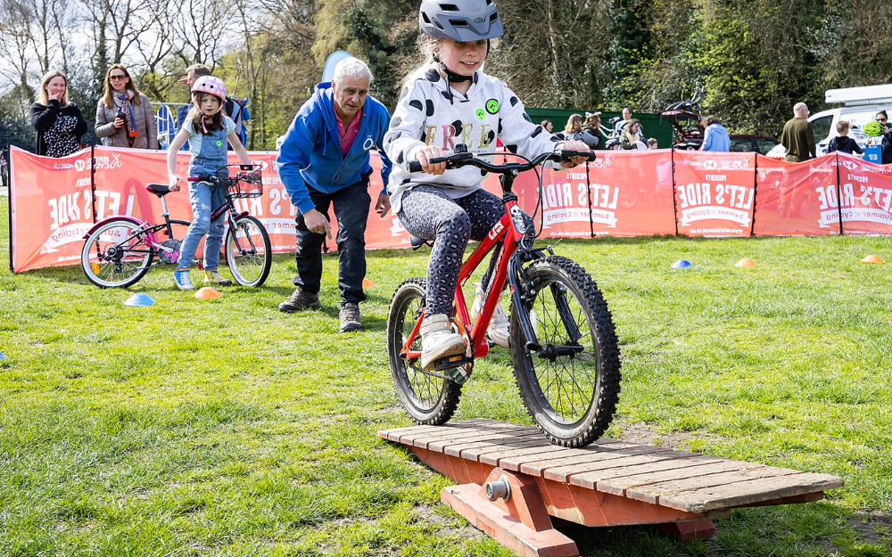 A girl in a cycle helmet rides over a small ramp while an instructor watches on