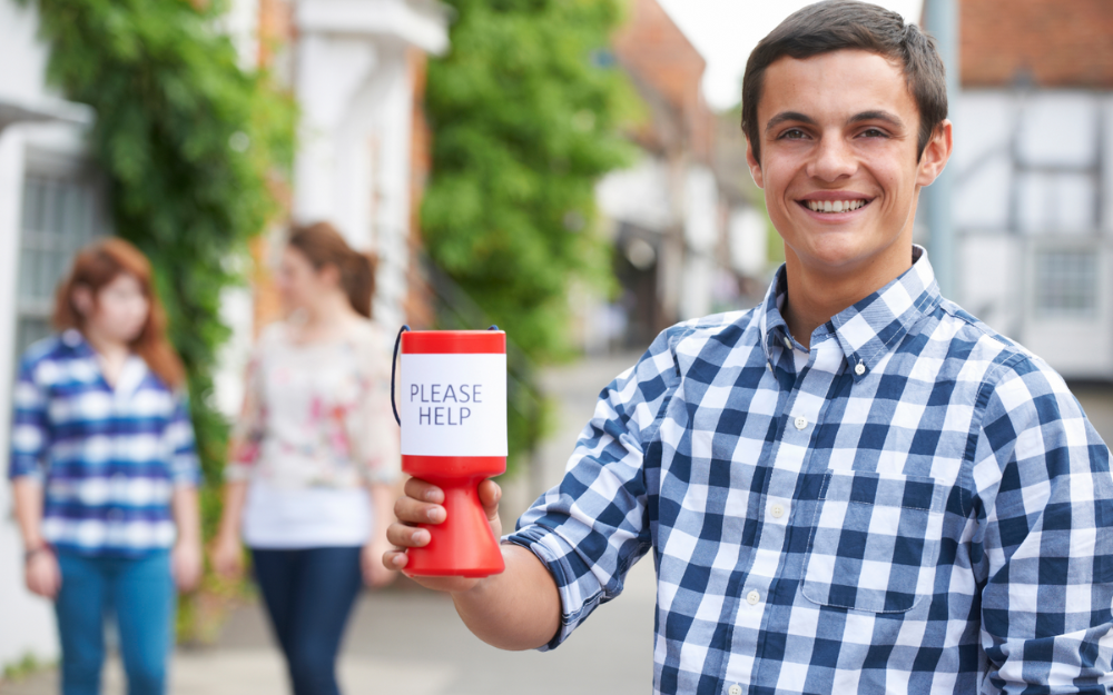A charity collector holding a colleciton tin, it has text on it which reads 'help me'