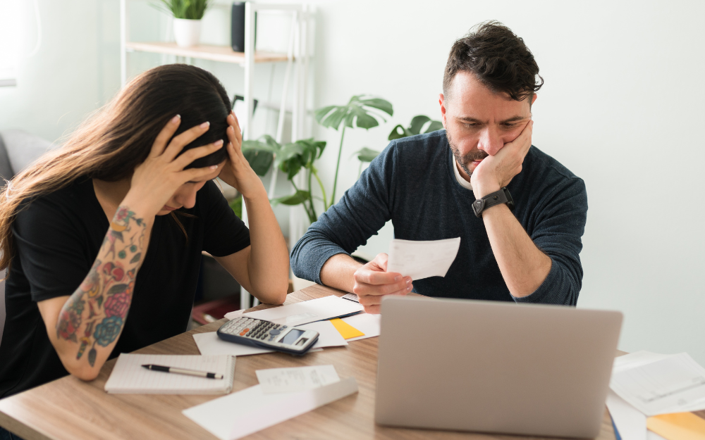 A couple sit around the table, looking distressed, with paperwork and calculators as they assess their finances
