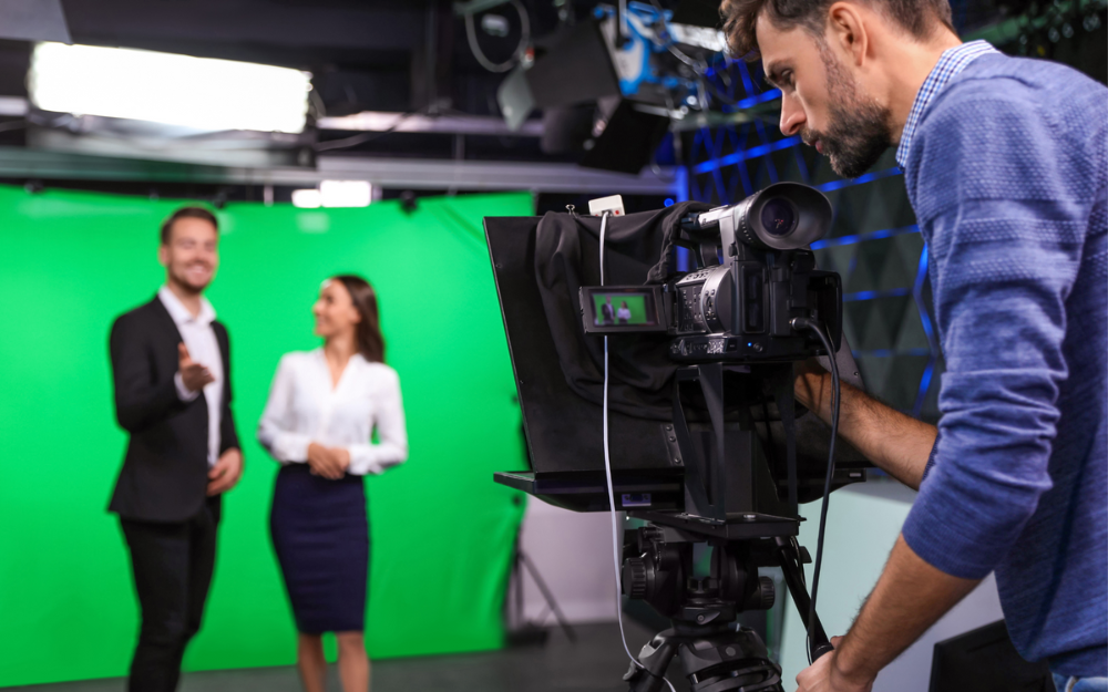 Two people in front of a green screen in a filming studio, being filmed by a camera operator