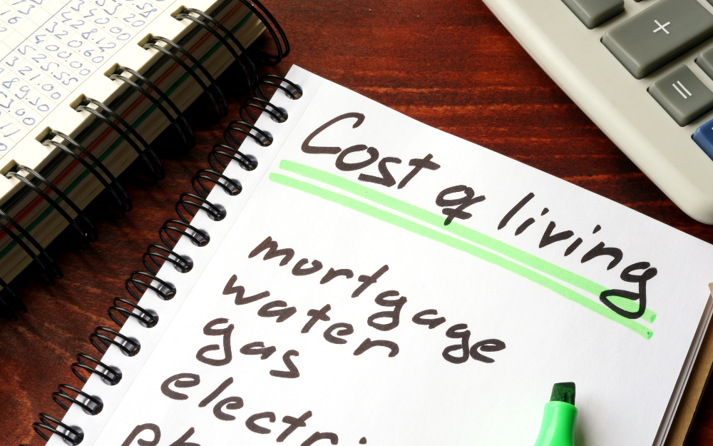 A notepad with cost of living written at the top, with a calculator and sheet with numbers next to it