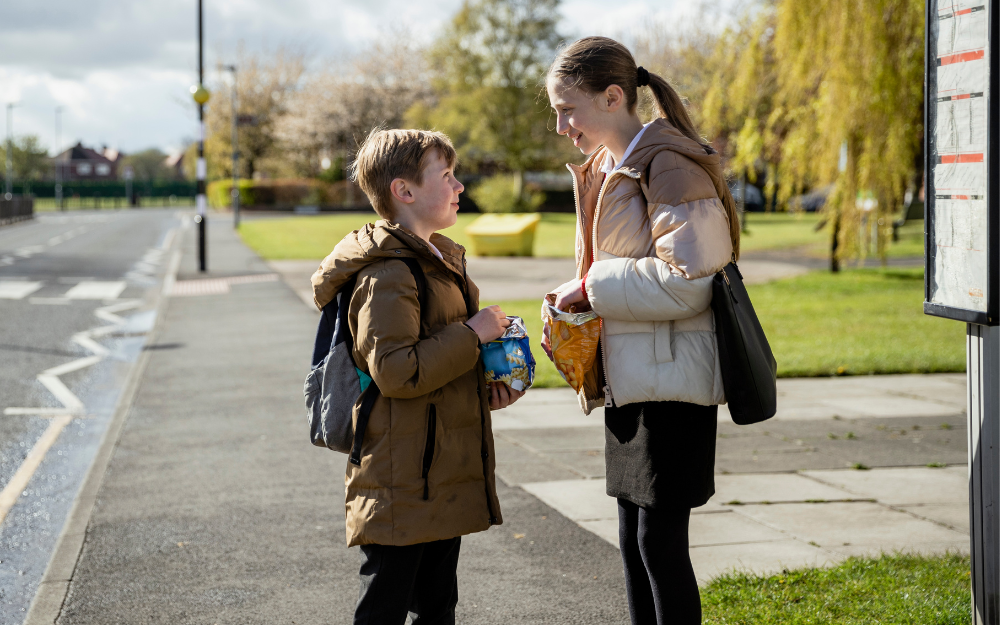 A boy and girl stand at a bus stop in their school uniforms