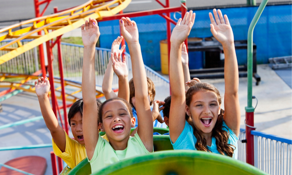 shot of children grinning and holding their arms up while riding a small rollercoaster