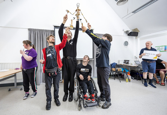 A group of young adults with learning disabilities, including a wheelchair user, jointly lift up a puppet in front of an audience