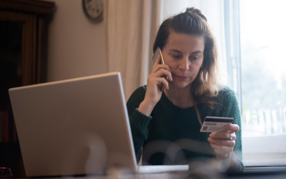 Woman sat in front of open laptop on the phone holding a debit card up to read