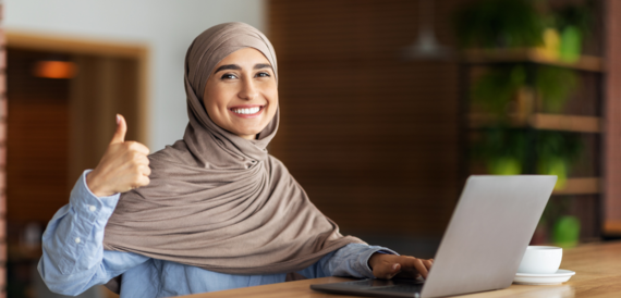 An adult female wearing a headscarf with an open laptop in front of her smiling and giving thumbs up to the camera