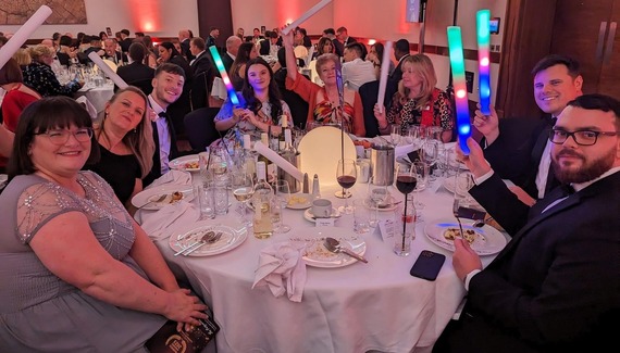 Happy people sitting around dinner tables at a large event, waving multi-coloured glowsticks