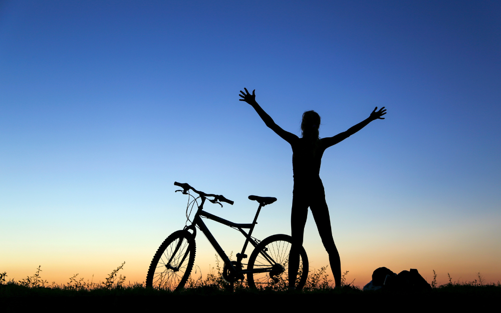 A woman stretches with joy next to her bike during the sunset