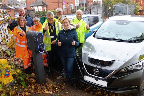 A group of men and women in high viz jackets stand around a new charger with a car plugged in and charging