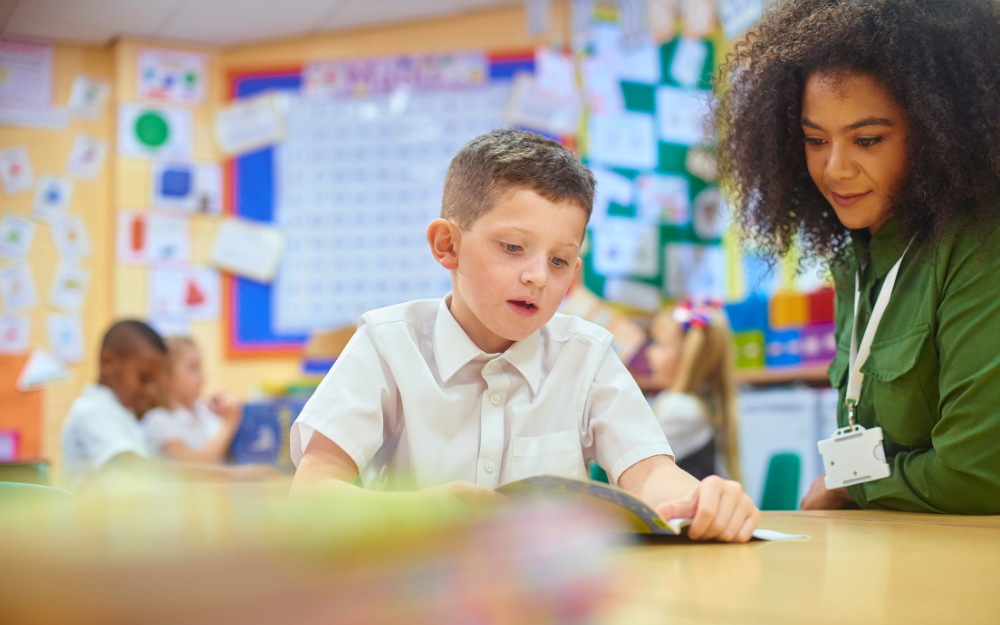 Primary school child looking at a book with a teacher looking on. In the background you can see other children and classroom displays