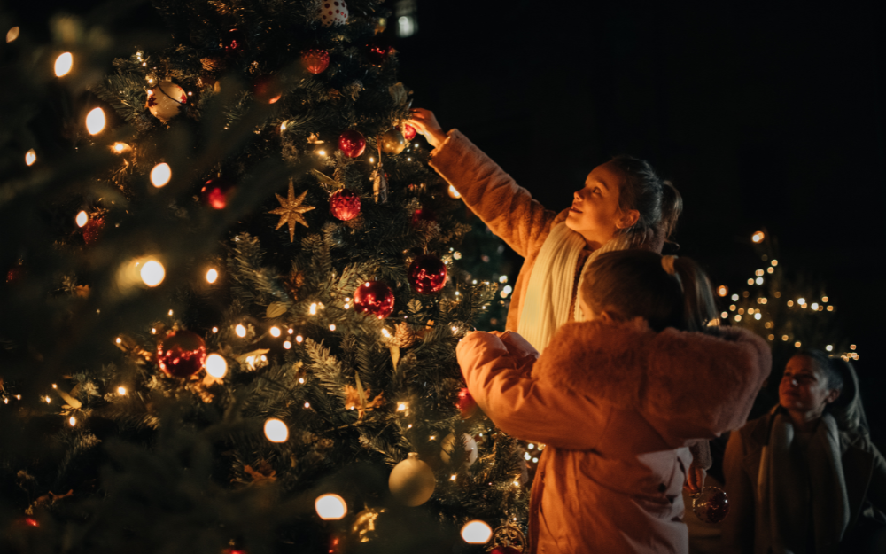 Two girls in warm coats place a decoration on an outdoor Christmas tree