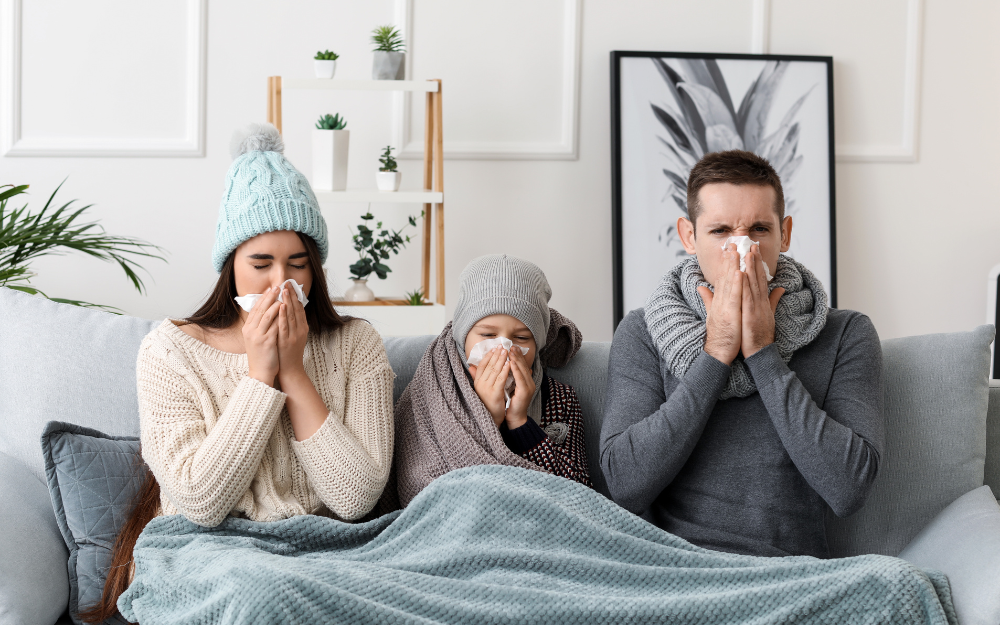 A family sat in thick jumpers, hats and scarves, under a blanket, blowing their noses