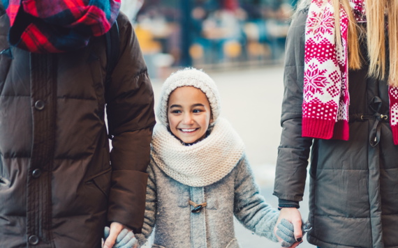 Two adults hold hands with a child, all are wrapped up warm in coats, scarves and gloves