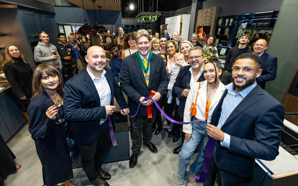 A large group of people stand in a kitchen showroom around a ribbon which they are preparing to cut