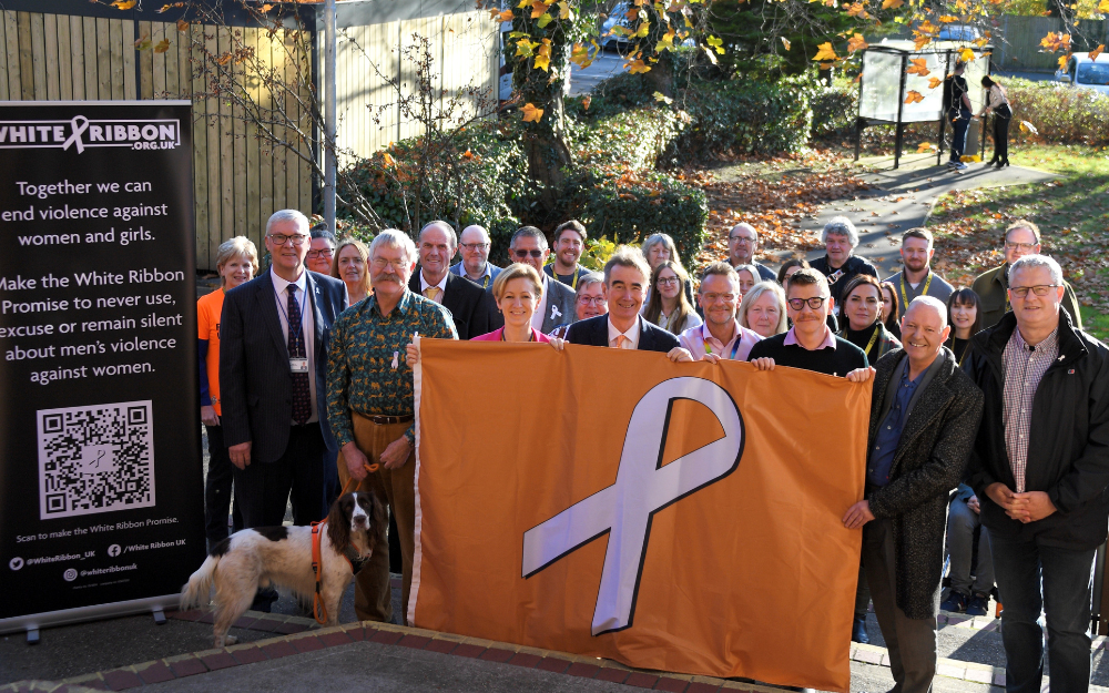 Councillors and officers stand outside the council building holding up the white ribbon flag, an orange flag with a white ribbon