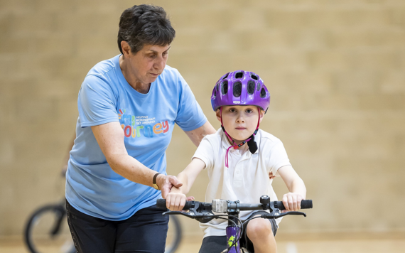 Jill holds the handlebars while a young boy wearing a purple helment practices riding